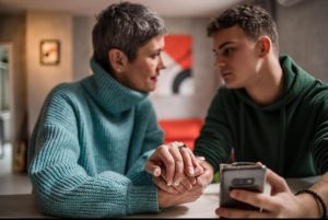A mother in her 40s sitting beside her teenage son, holding his hand, with a calm and supportive expression.
